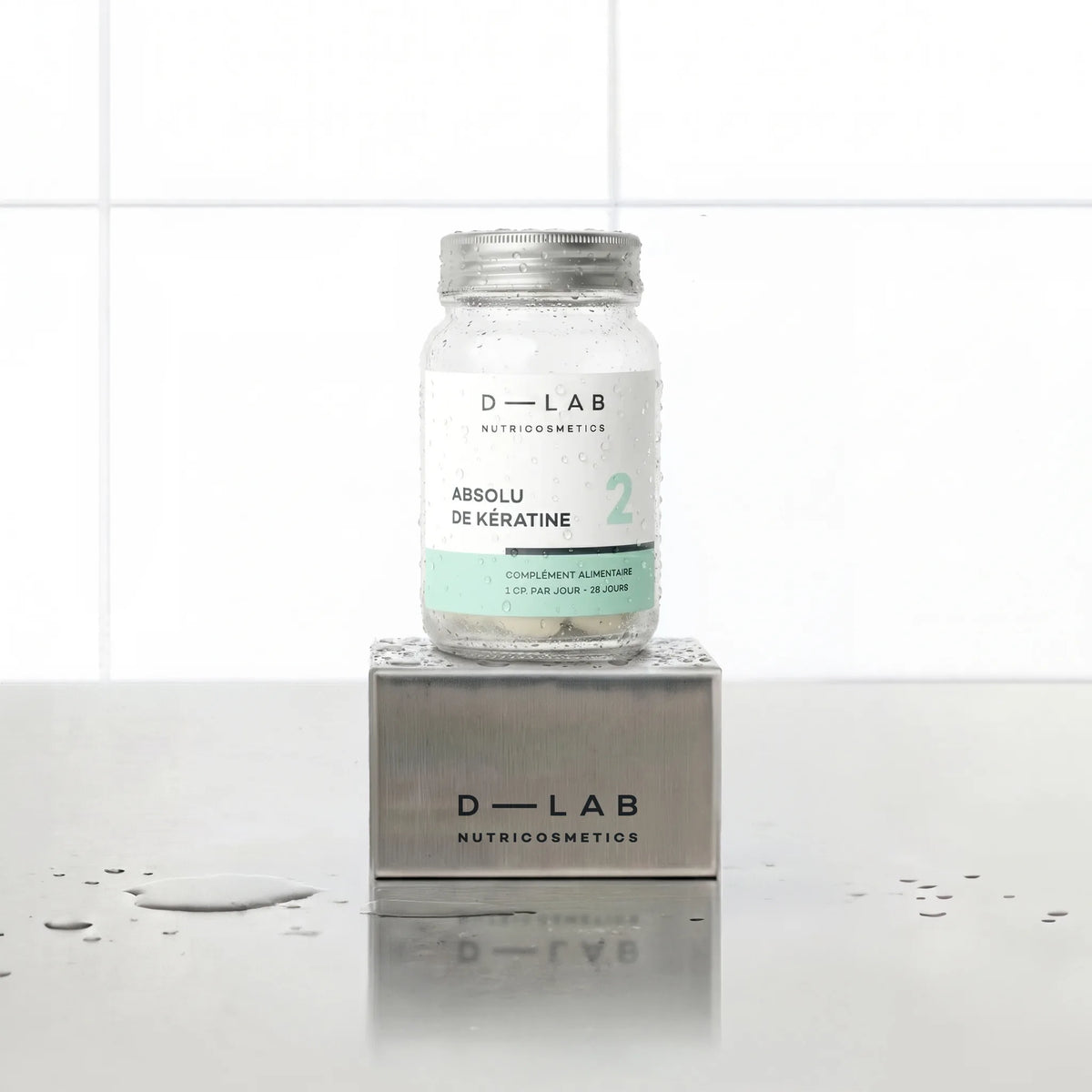 Absolu de Kératine D-LAB Nutricosmetics cure 1 mois, flacon en verre de gélules posé sur un socle inox brossé, fond carrelage blanc et gouttes d'eau