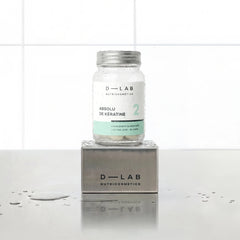 Absolu de Kératine D-LAB Nutricosmetics cure 1 mois, flacon en verre de gélules posé sur un socle inox brossé, fond carrelage blanc et gouttes d'eau