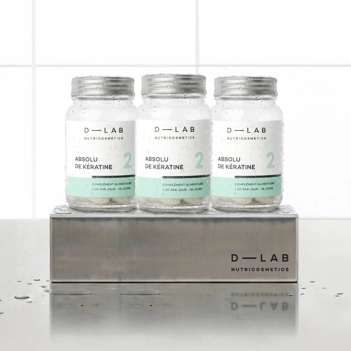 Absolu de Kératine D-LAB Nutricosmetics cure 3 mois, trois flacons en verre de gélules alignés sur un socle inox brossé, décor carrelage blanc