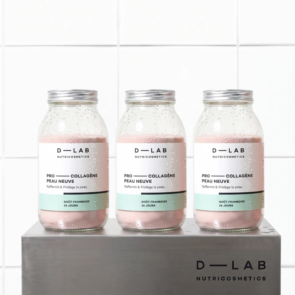 Cure 3 mois Pro-Collagène Peau Neuve goût framboise D-LAB, trois flacons en verre de poudre rose sur socle en métal brossé, fond carrelage blanc