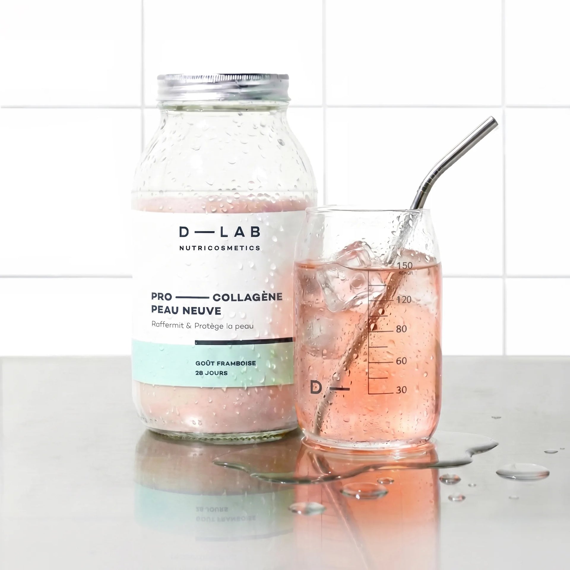 Flacon en verre Pro-Collagène Peau Neuve goût framboise D-LAB et verre glacé de boisson rosée avec glaçons et paille inox, plan de travail carrelé blanc