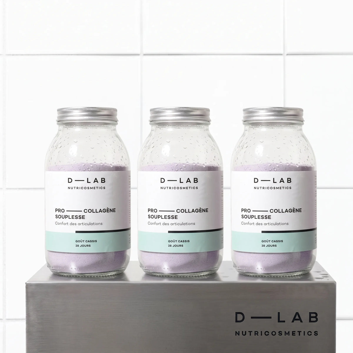 Cure 3 mois Pro-Collagène Souplesse goût cassis D-LAB, trois flacons en verre de poudre mauve sur socle en métal brossé, fond carrelage blanc