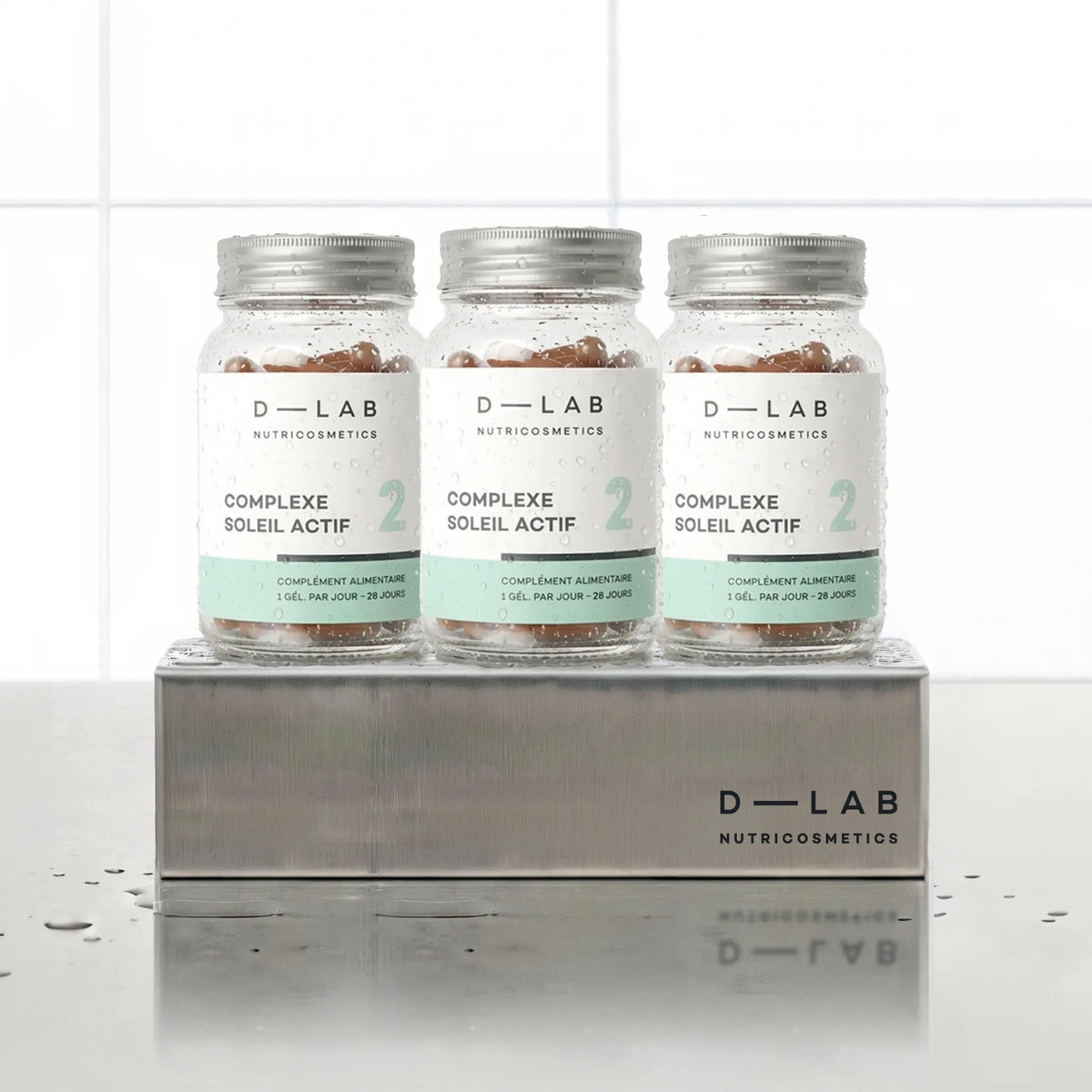 Complexe Soleil Actif D-LAB Nutricosmetics, cure 3 mois : 3 flacons de gélules alignés sur un socle inox brossé, fond carrelage blanc