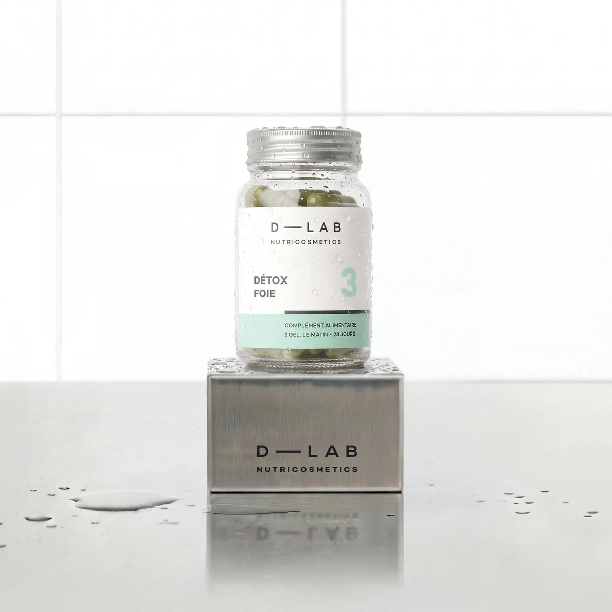 Détox Foie D-LAB Nutricosmetics, cure 1 mois : flacon en verre de gélules posé sur un socle inox brossé, fond carrelage blanc