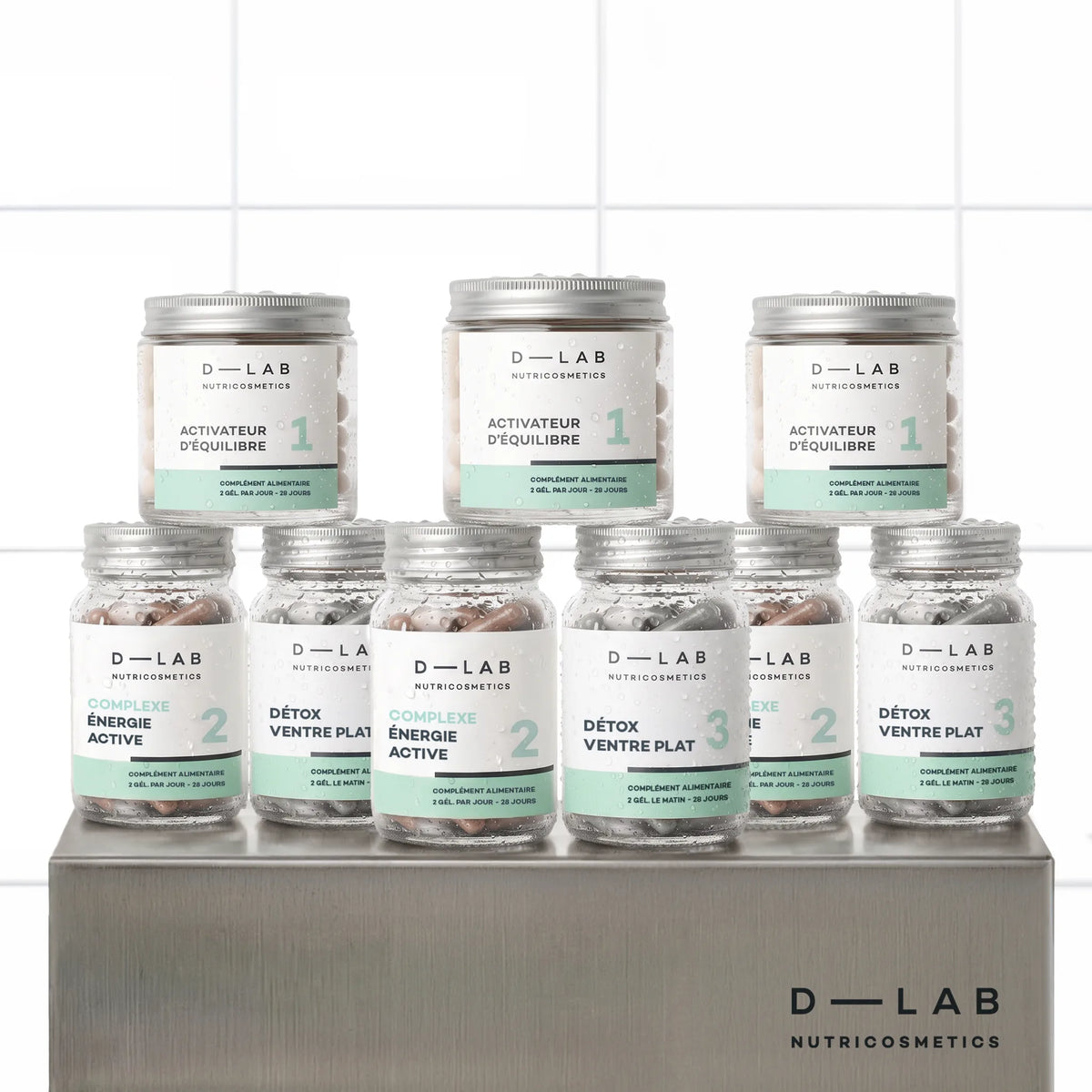 Routine Ventre Plat D-LAB cure 3 mois, neuf pots du programme en trois exemplaires alignés sur un socle inox brossé, fond carrelage blanc