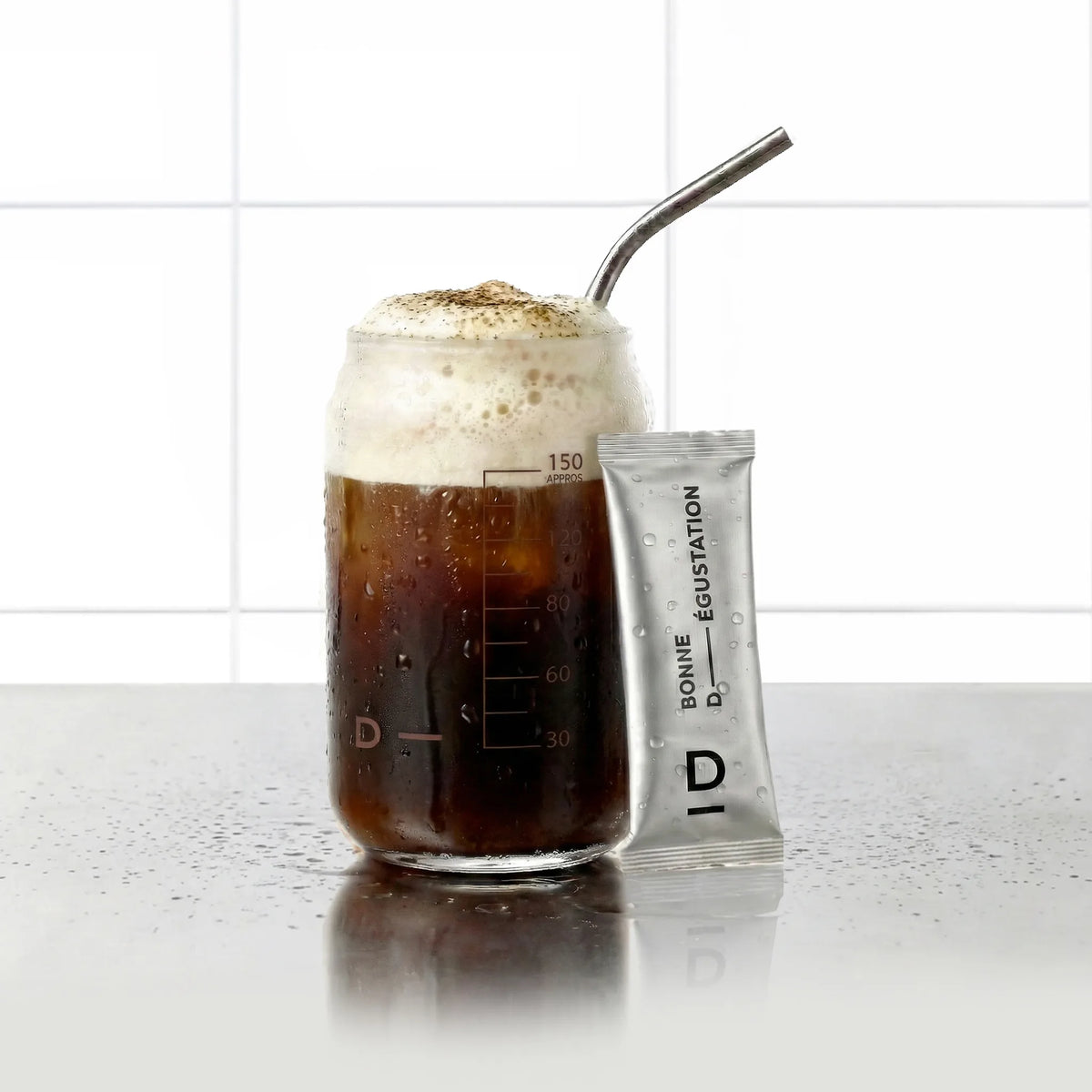 Sticks Brûle Graisses Café D-LAB, stick de poudre posé contre un verre de boisson café mousseuse avec paille inox, fond carrelage blanc