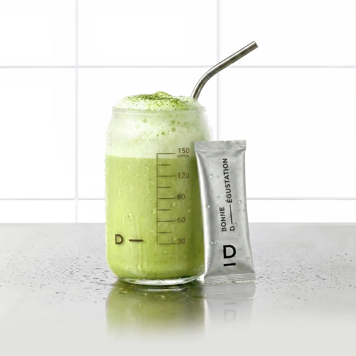 Sticks Peau Neuve Matcha D-LAB, stick de poudre posé contre un verre de boisson matcha mousseuse verte, fond carrelage blanc